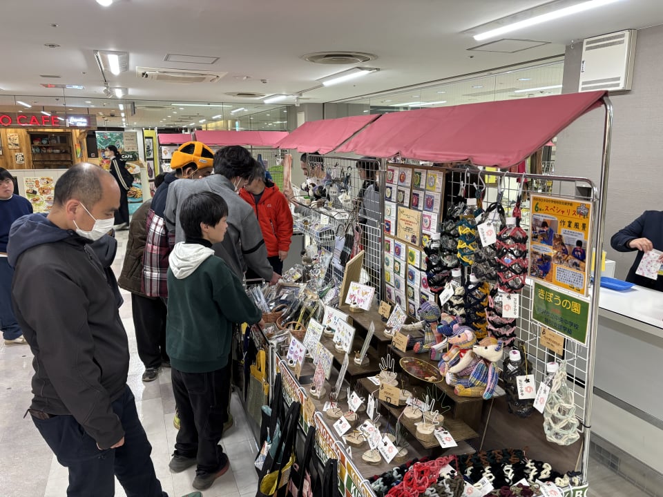 手しごと展示販売会の写真。きぼうの園の出店ブースに2名の方が視線を注いで作品を見つめている。